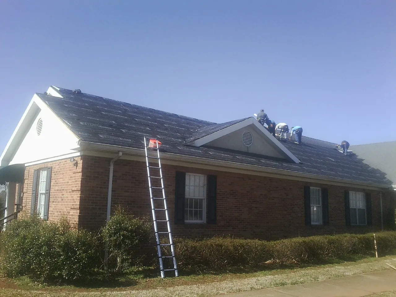 Crew tearing shingles off a brick ranch roof with a ladder set — Metal Roof Repair work by our crew in East Hickory