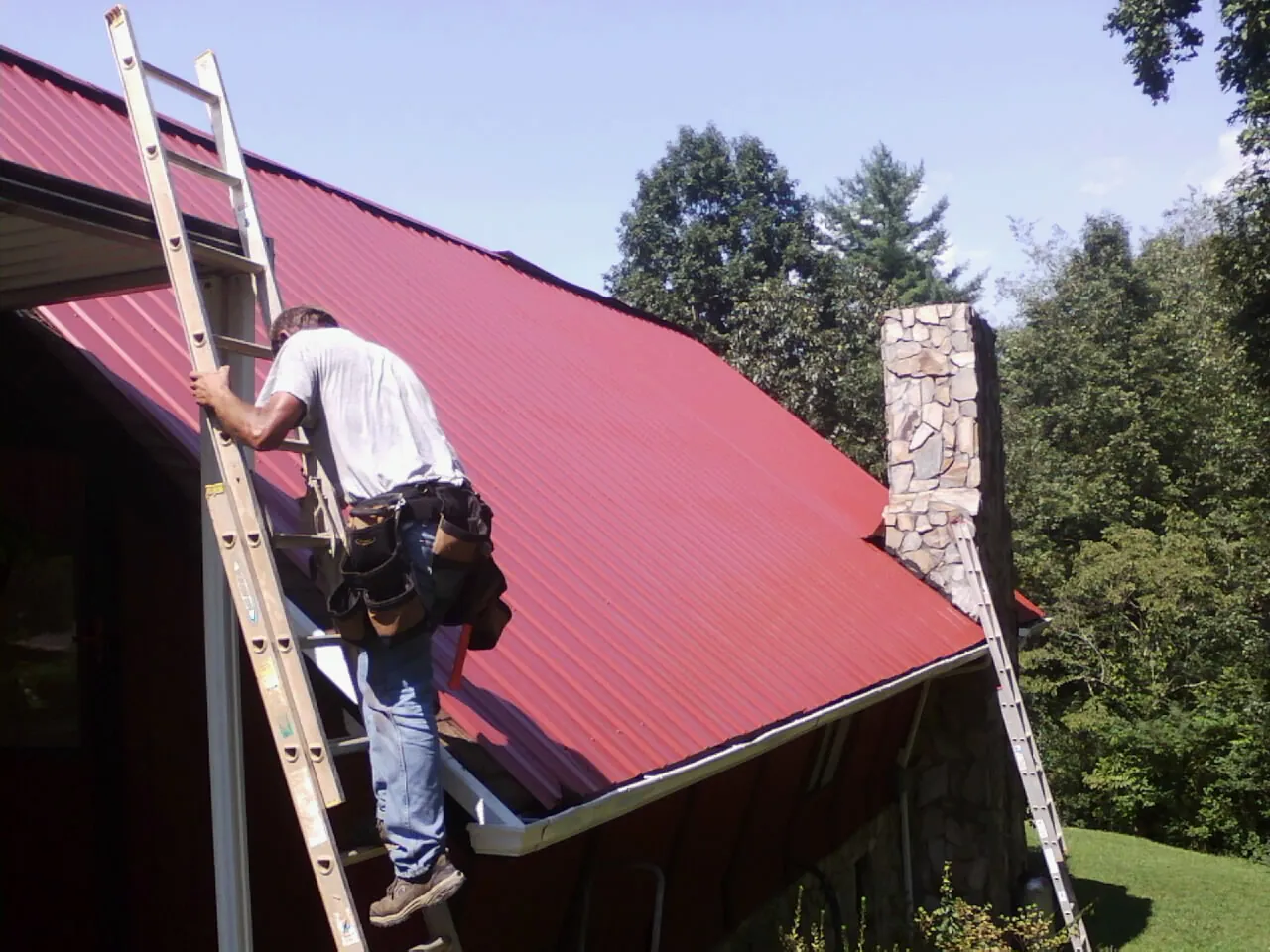 Roofer on a ladder beside a red metal roof and stone chimney — Soffit & Fascia Repair by Roofing Brothers in East Hickory